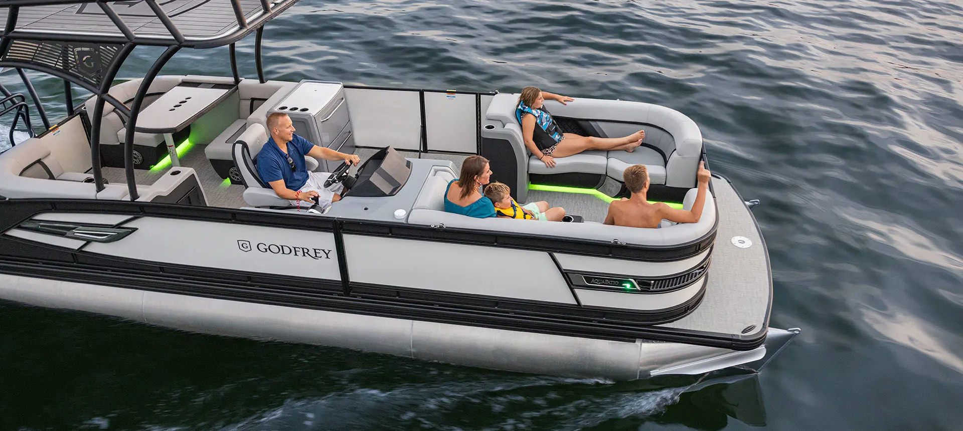 Aerial side view of a Godfrey AquaPatio Entertainment pontoon boat cruising on a lake.
