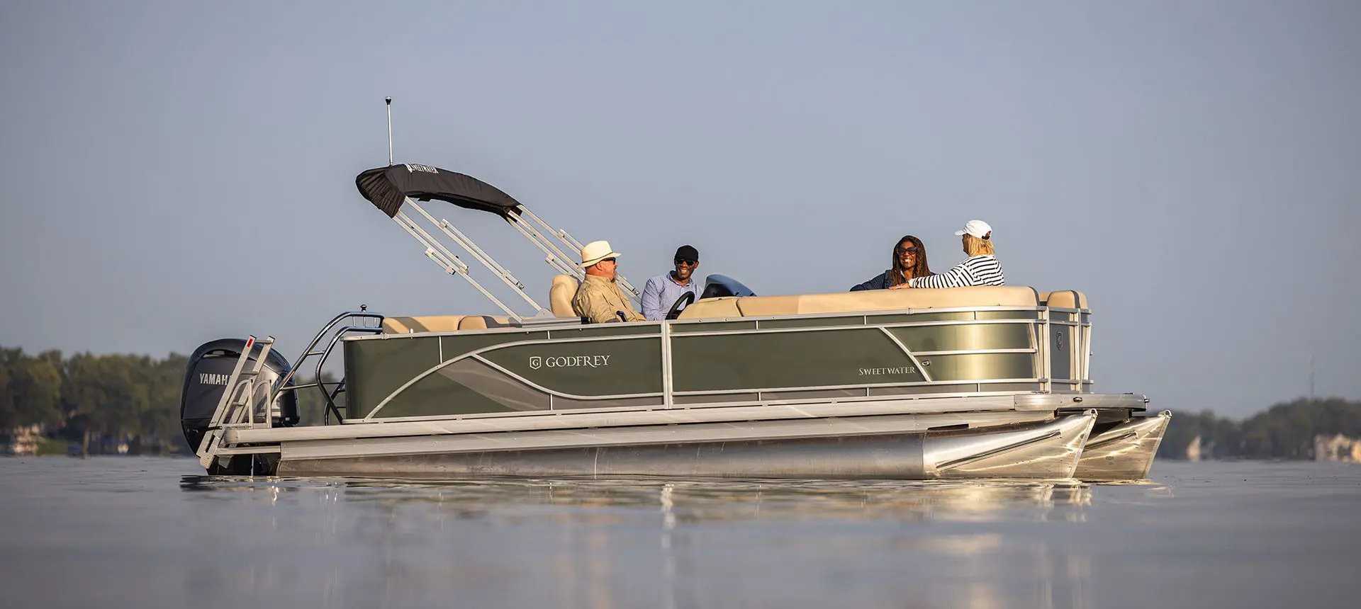 Side profile of a Godfrey Sweetwater Cruise pontoon boat floating on the lake with 4 people on board.