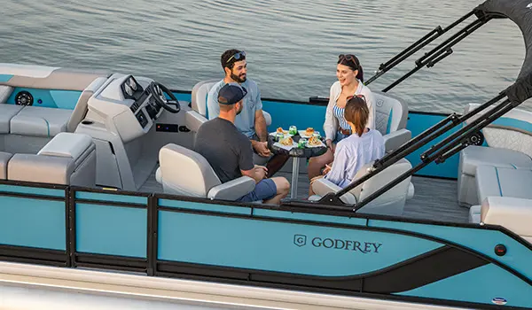 Side profile of a Godfrey Sweetwater Entertainment pontoon boat floating on a lake at sunset.