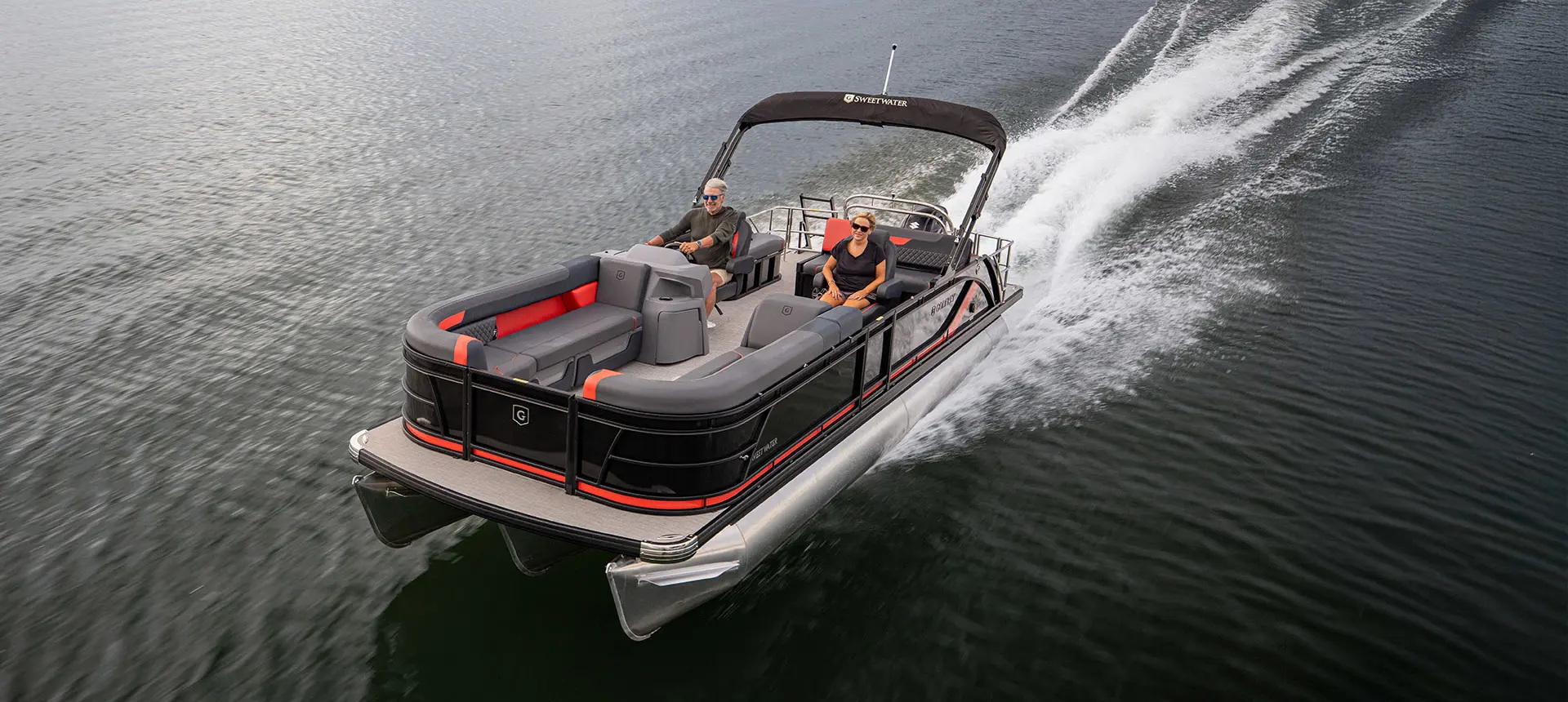 Bow focused aerial view of a couple cruising the lake on a Godfrey Sweetwater Lounge pontoon boat.