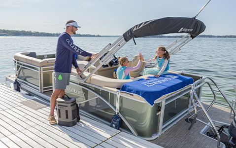 Girls high-fiving on the back of a pontoon boat while wearing Godfrey life jackets while sitting near a Godfrey towel,.