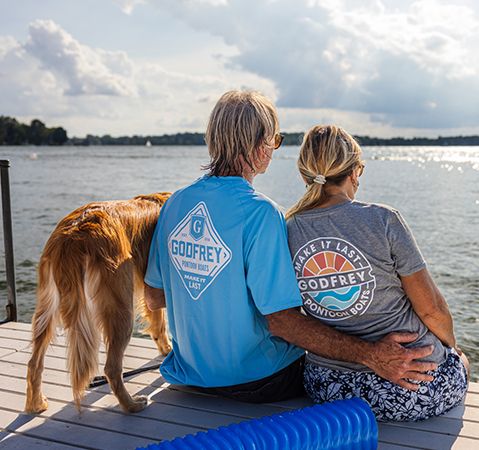 Couple and their dog sitting on a dock, both wearing Godfrey Pontoon Boats branded t-shirt.