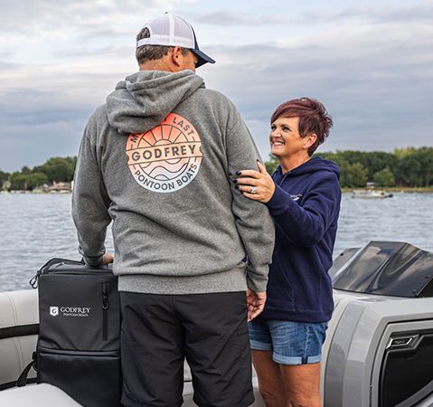 Man wearing a gray Godfrey Pontoon Boats hoodie and her partner smiling at him on a pontoon boat.