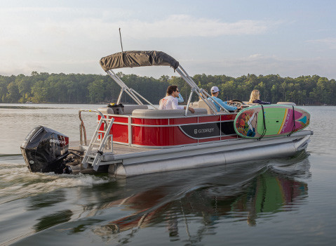 Side-view of a Godfrey pontoon boat with a surf board attached to the side with toolless boat accessory attachment system. 