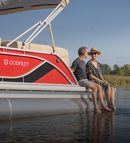 Man and woman sitting on back of a Godfrey Pontoon Boat with their toes in the water. 