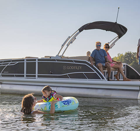 A couple sitting on the jump seat of a Godfrey pontoon boat with the Cruise layout watching 2 people swimming in the water.