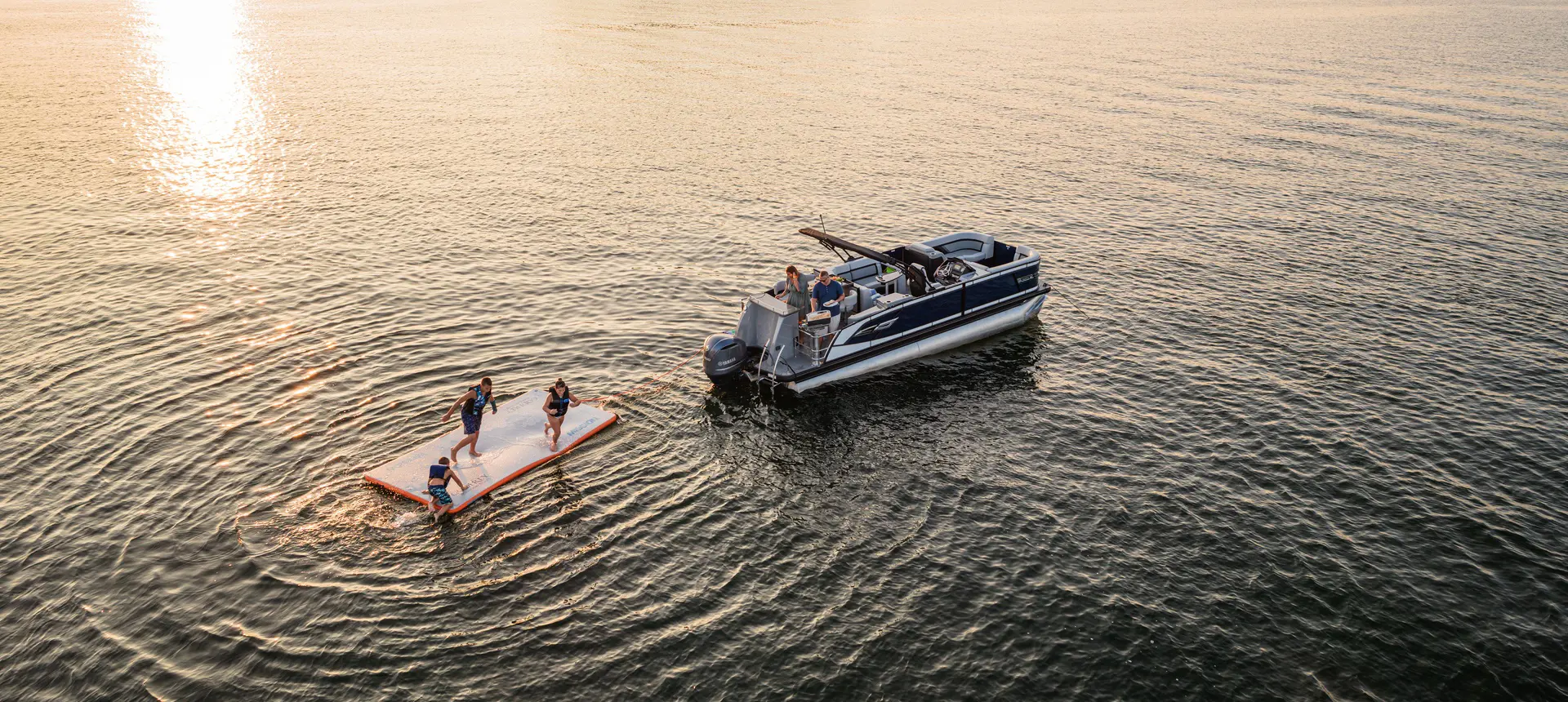 Aerial view of kids jumping off a float hooked to a Godfrey AquaPatio pontoon boat on a lake.
