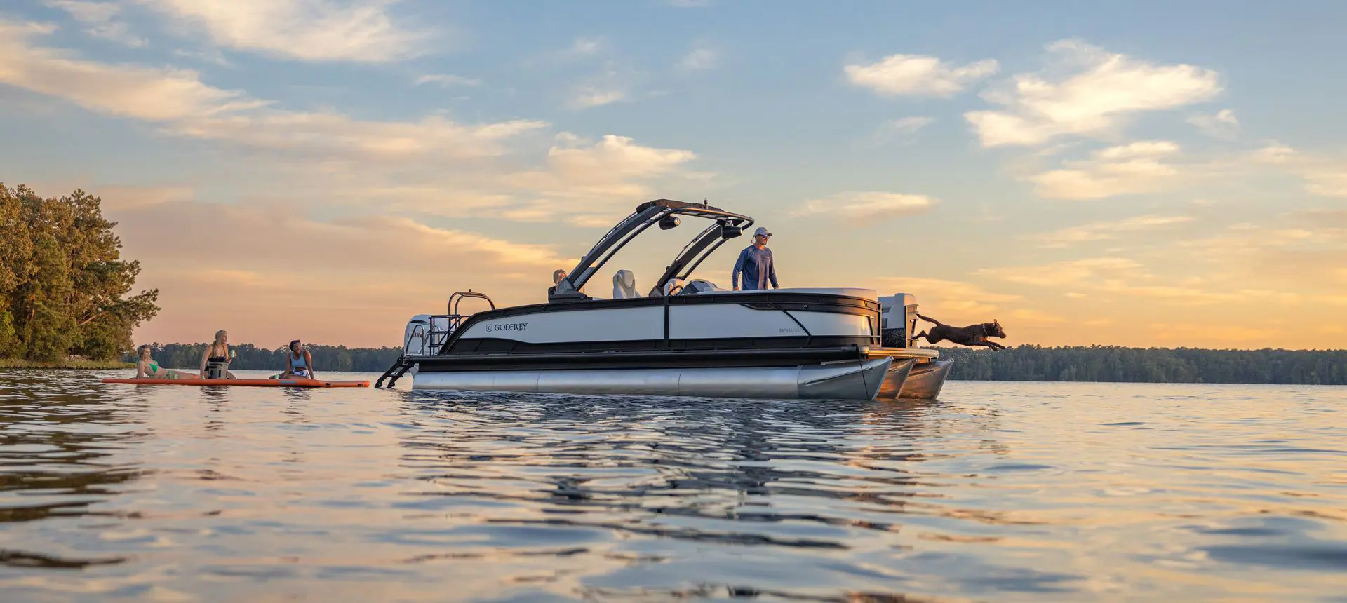 Side profile of a Godfrey Monaco pontoon boat floating on a lake pulling a floating pad with people on it.