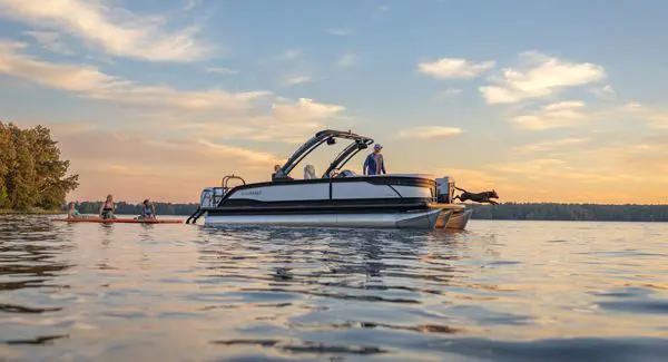 Side profile of a Godfrey Monaco pontoon boat floating on a lake pulling a floating pad with people on it.