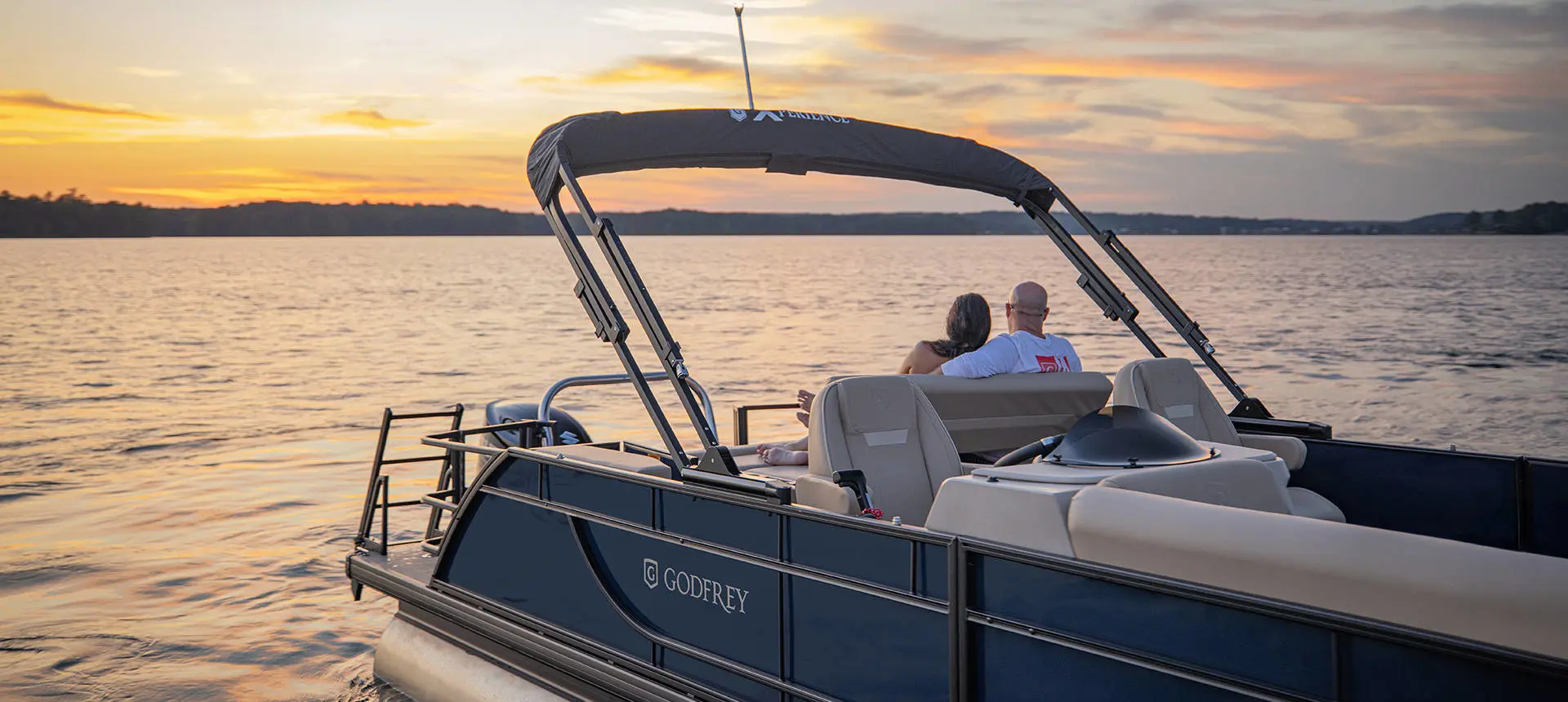 A couple relaxing on the stern seating of a Godfrey Xperience pontoon boat while floating on a lake.