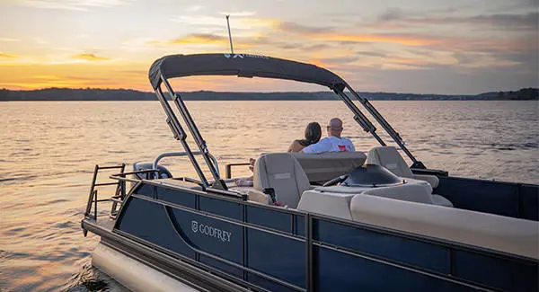 A couple relaxing on the stern seating of a Godfrey Xperience pontoon boat while floating on a lake.