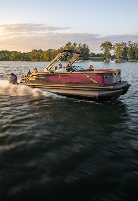 Godfrey AquaPatio luxury pontoon boat carrying passengers across a lake in warm evening light.