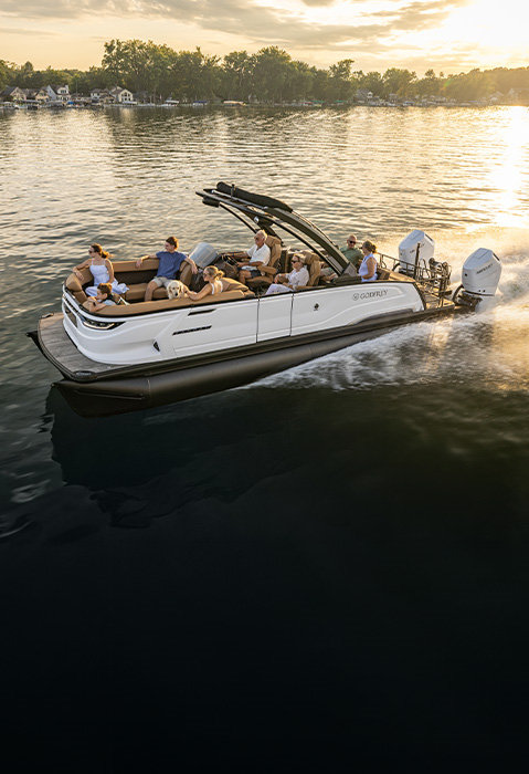 White Godfrey Sanpan luxury pontoon boat with twin outboard engines carrying passengers and a dog across a sunlit lake.
