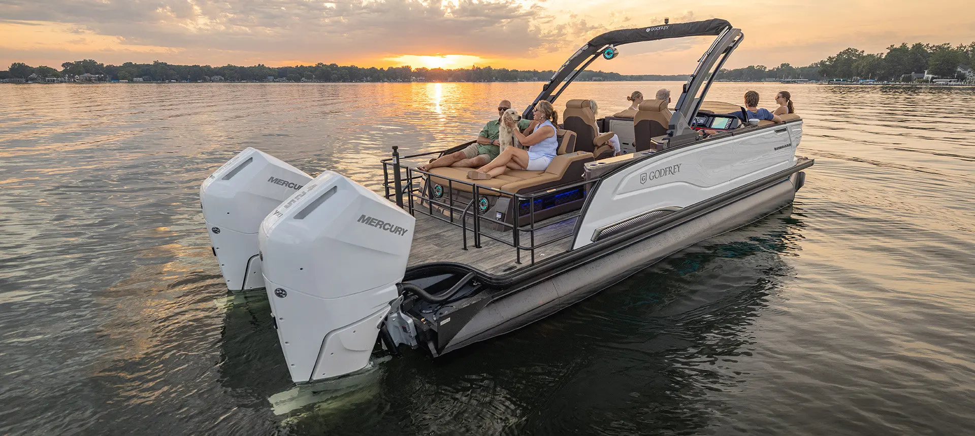 2 people sitting with their dog on the stern flip lounge seat on a Godfrey Sanpan pontoon boat at sunset.