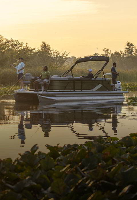 Passengers fish from a Godfrey Sweetwater pontoon boat on a calm, plant-lined lake in warm evening light.