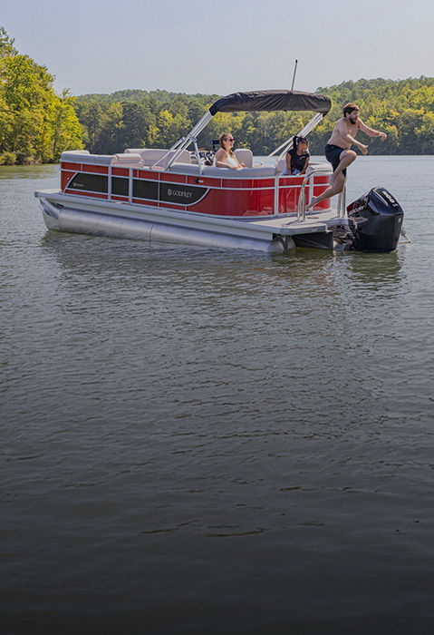 Two people jump from a red Godfrey Xperience pontoon boat as two passengers remain aboard on a forest-lined lake.