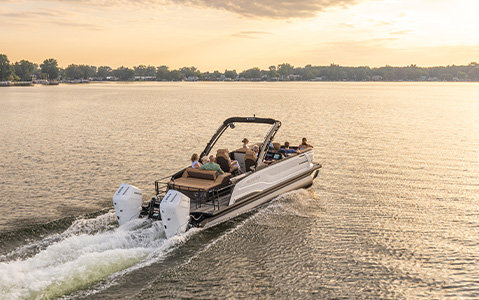 Godfrey AquaPatio pontoon boat with twin outboard engines carrying passengers across a lake at sunset.
