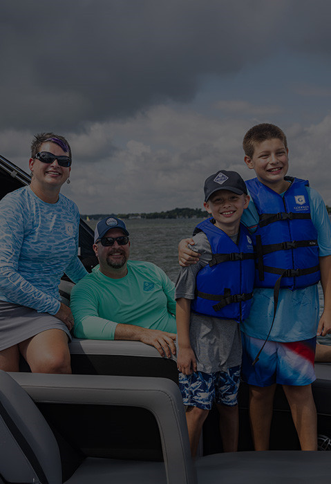 Family smiles on the back of a Godfrey Pontoon boat, wearing Godfrey Pontoon accessories and apparel. 