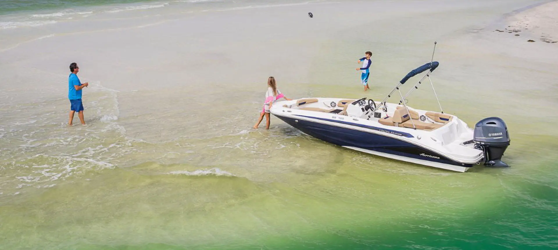 Dad and son playing catch in front of a Hurricane SunDeck Sport 185 OB deck boat on the beach.