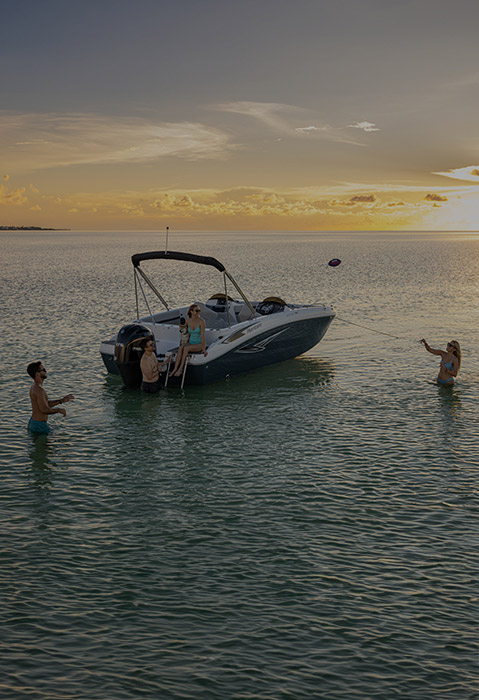 A Hurricane Deck Boat is anchored in calm water during sunset, with people swimming and playing catch with a football around the boat.