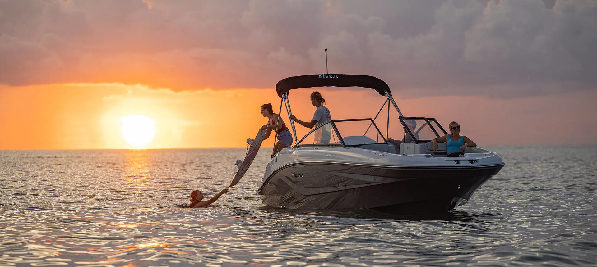 A person on a Hurricane SunDeck OB deck boat handing a wakeboard to a person wearing a life jacket floating in the ocean.