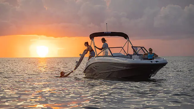 A person on a Hurricane SunDeck OB deck boat handing a wakeboard to a person wearing a life jacket floating in the ocean.