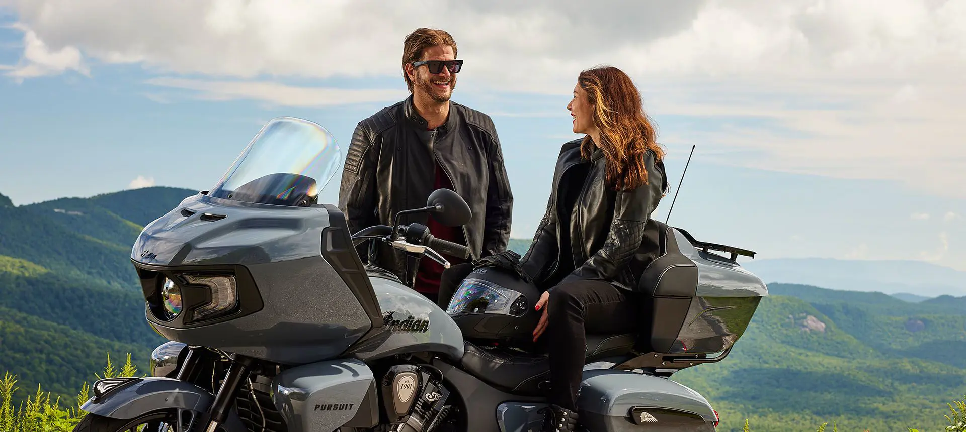 a woman sitting on an indian pursuit motorcycle while a man stands nearby