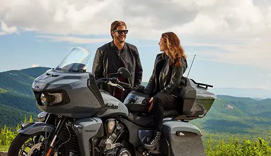 a woman sitting on an indian pursuit motorcycle while a man stands nearby
