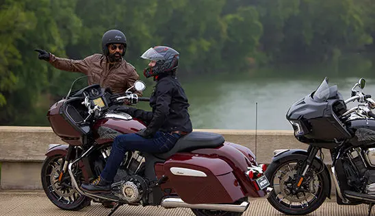 person sitting on a maroon metallic indian challenger limited motorcycle while a man nearby provides direction