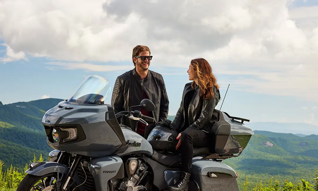a woman sitting on an indian pursuit motorcycle while a man stands nearby smiling