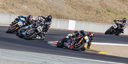 Tyler O’Hara leads a group of motorcycle racers in full gear lean into a sharp turn on a race track.