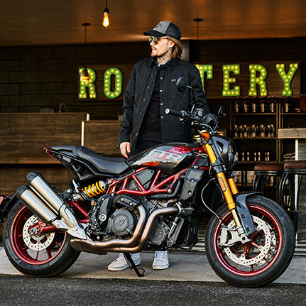 Man standing behind the chiseled and muscular Indian FTR R Carbon standard-style motorcycle.