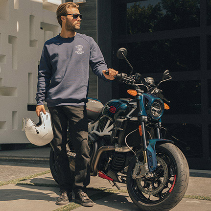 Man standing near an Indian FTR x 100% R Carbon standard-style motorcycle while a woman watches in the background.