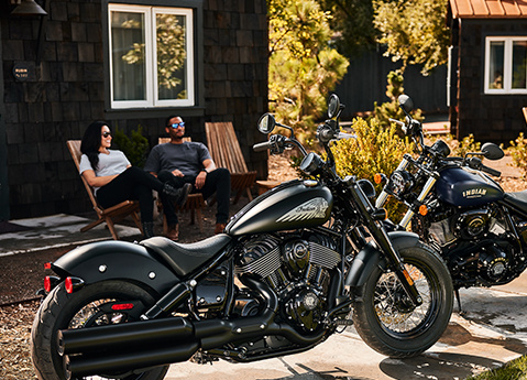 Side profile of a 2025 Indian Chief Bobber Dark Horse cruiser motorcycle with 2 people sitting in the background.