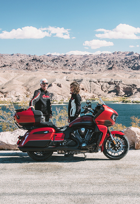 A couple standing beside a 2025 Indian Pursuit Dark Horse motorcycle near a lake.