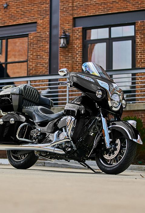 Side/front profile of a 2025 Indian Roadmaster touring motorcycle parked in front of a brick building.