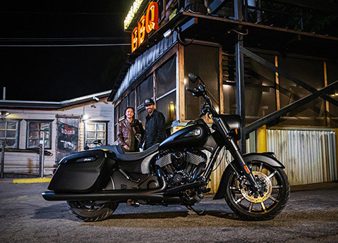Two guys smiling toward a 2026 Indian Springfield Dark Horse bagger motorcycle while outside a business at night.