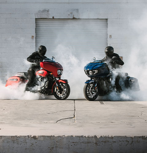 Two motorcyclists on red and blue Indian Motorcycles perform burnouts, creating smoke against a gray wall.