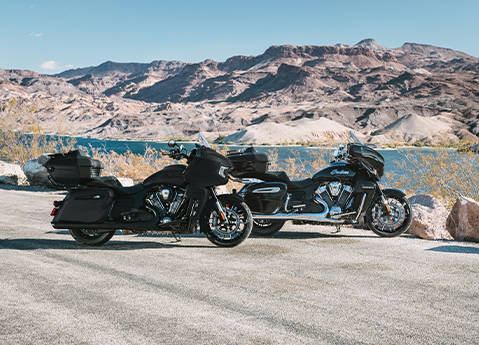 Side profile of an Indian Pursuit Limited touring motorcycle and a Indian Pursuit Dark Horse touring motorcycle parked out in the sunlight.