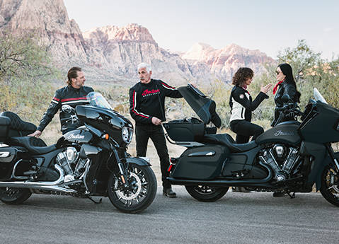 A man grabbing gear from the storage trunk on a Indian Pursuit Dark Horse touring motorcycle while 3 other people are nearby. 