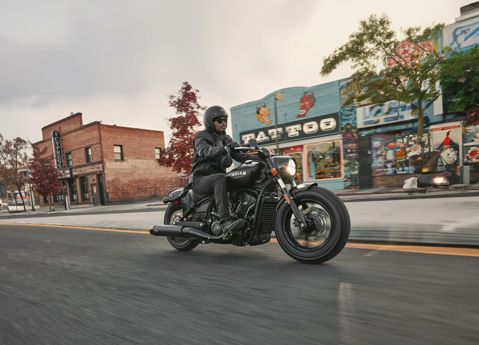 A person in a helmet riding an Indian Scout mid-size cruiser motorcycle through a city.