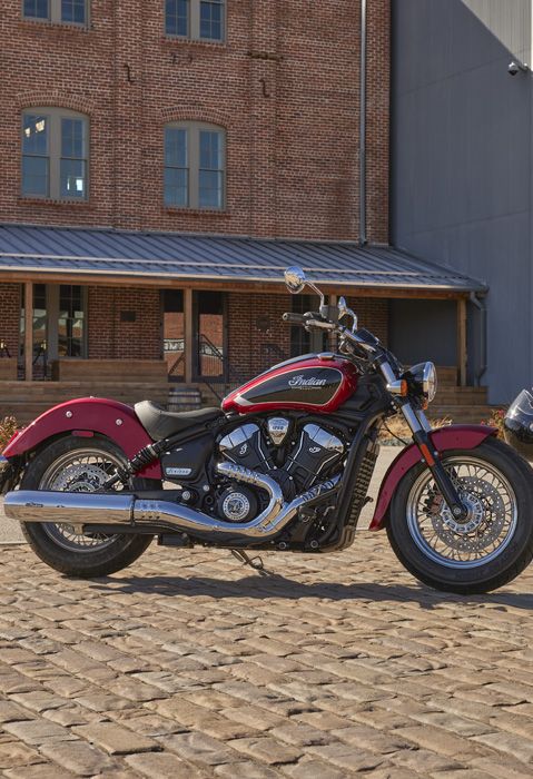 A woman carrying a helmet and walking toward an Indian Scout cruiser motorcycle on a cobblestone street. 