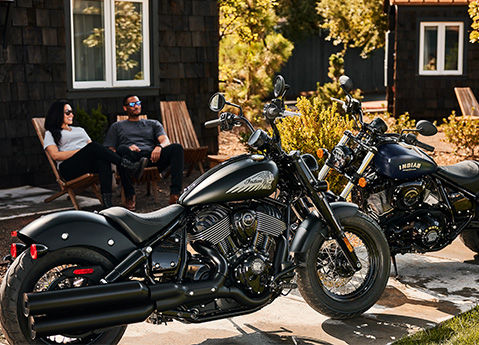 2 people sitting in chairs behind their parked 2026 Indian Chief Bobber cruiser motorcycles.