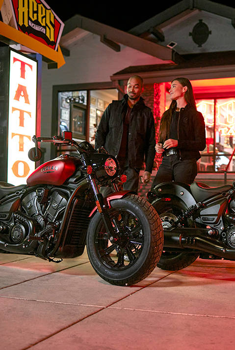 A man and a woman standing behind their 2026 Indian Scout Bobber cruiser motorcycles outside of a tattoo parlor.