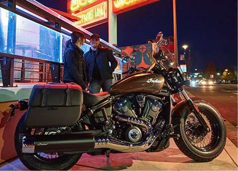 2 people standing behind a 2026 Indian Super Scout cruiser motorcycle parked on the side of the road at night.