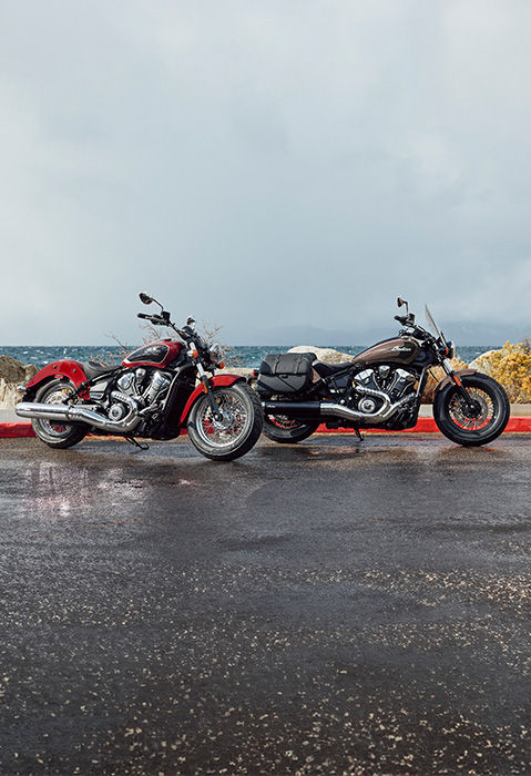 Two 2026 Indian Super Scout cruiser motorcycles parked on the side of the road with a lake in the background.