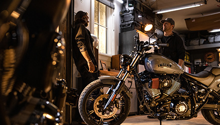 A man and a woman chatting inside a garage, an Indian motorcycle is parked in front of them
