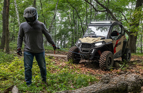 A person pulling out the winch from the front end of a Polaris RZR Trail side-by-side while in the forest.