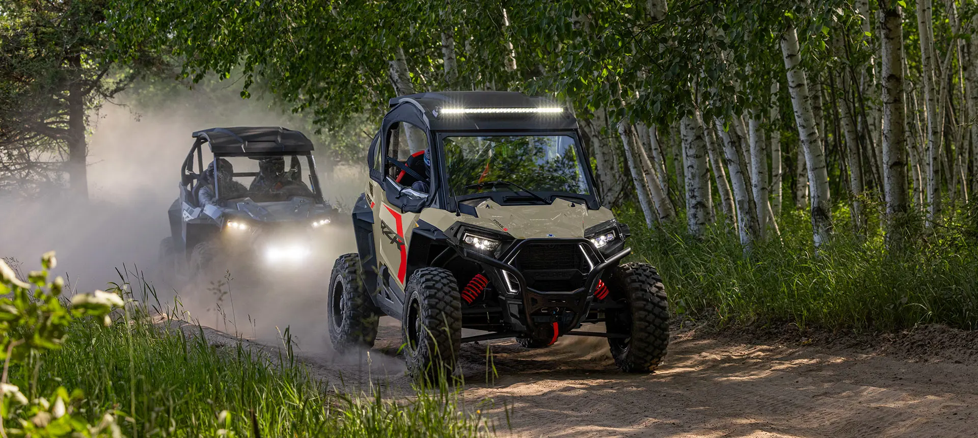 Two Polaris RZR Trail side-by-sides driving on a trail through the forest.