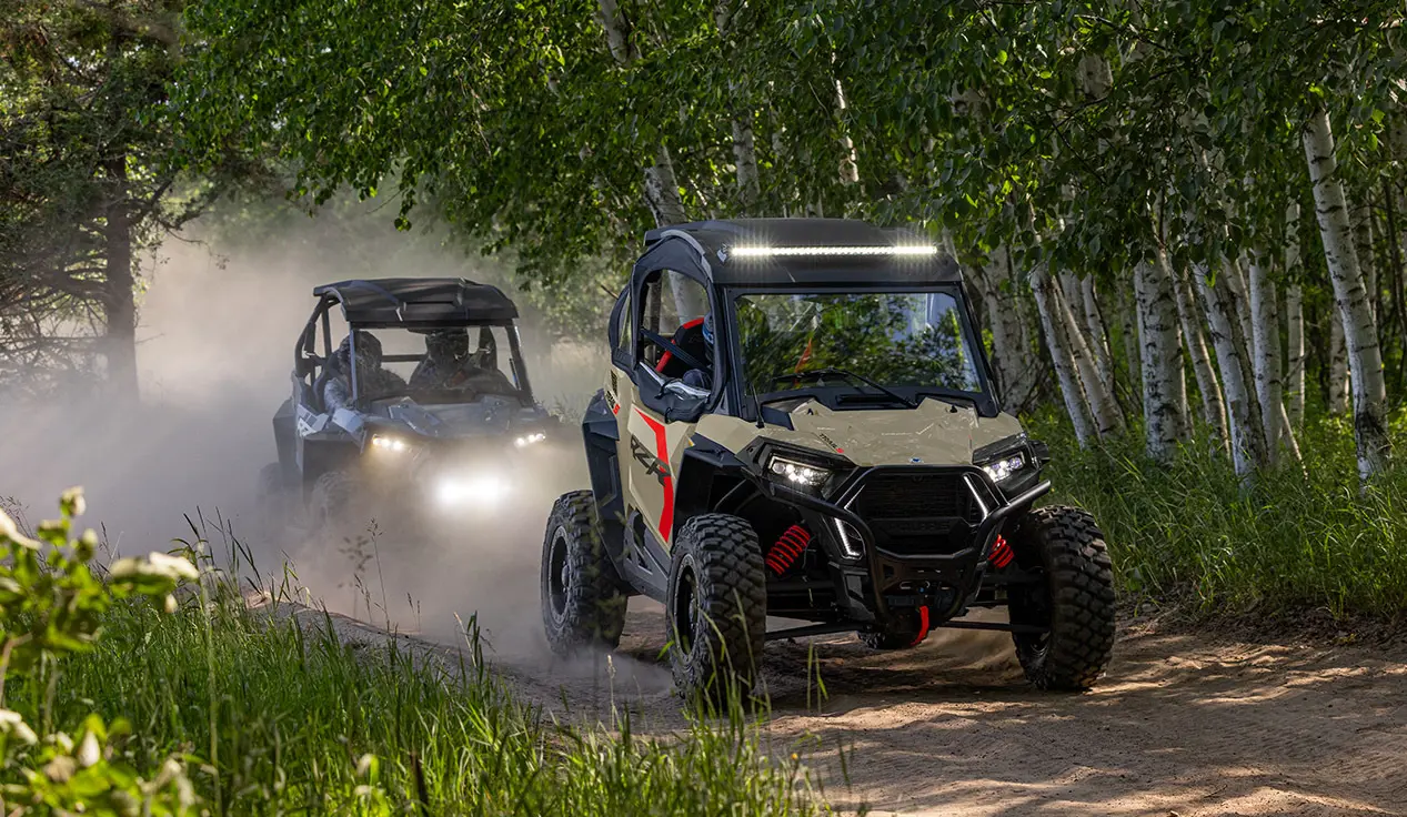 Two Polaris RZR Trail side-by-sides driving on a trail through the forest.